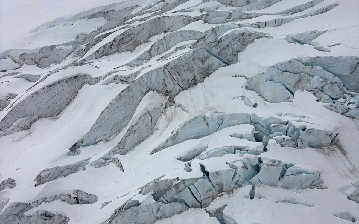 Deep crevasses running across a white glacier, seen straight down from the air