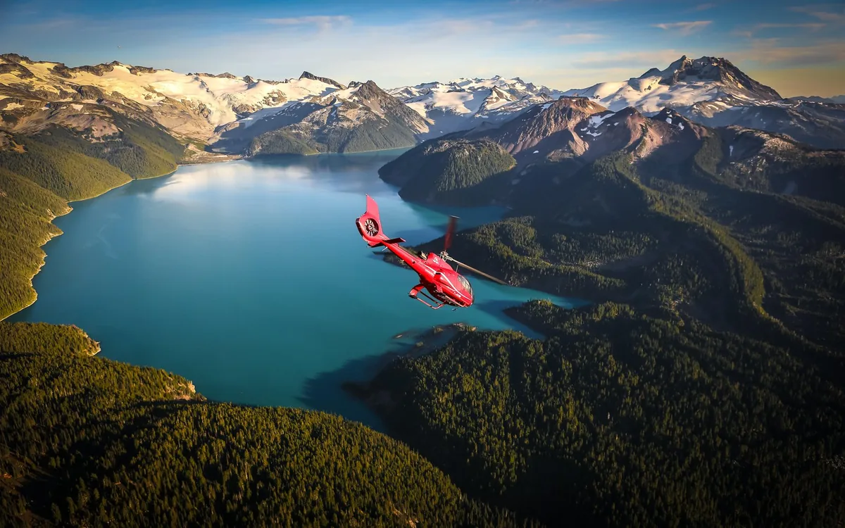 A red helicopter flying above a turquoise lake ringed by forest