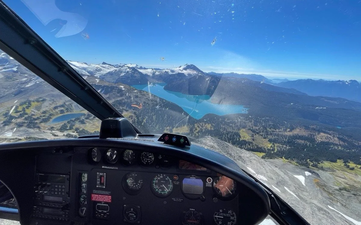 The view forward over a helicopter instrument panel to a turquoise lake and a line of snow-covered peaks