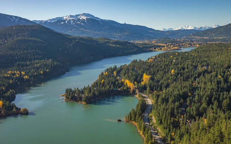 An aerial view along a lake and a valley road through forest, with snowy peaks behind