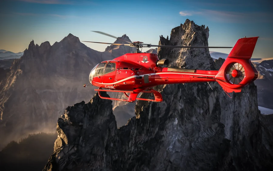 A red helicopter turning past jagged rock spires above a valley at sunrise