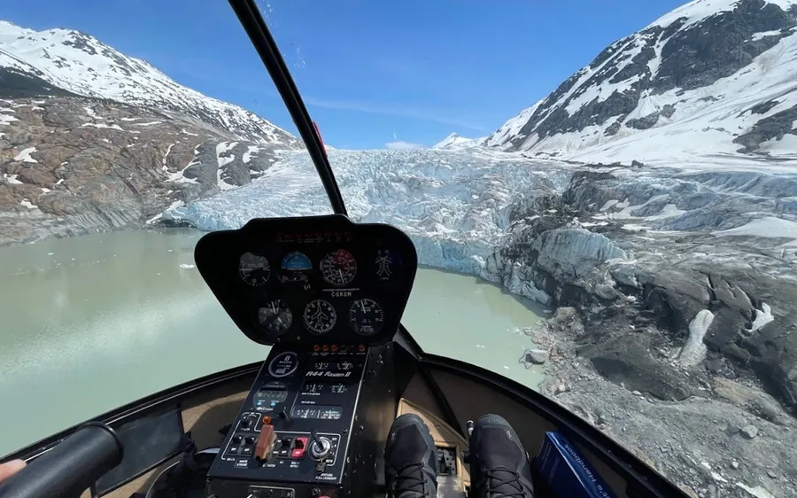 The view from a helicopter cockpit past the instruments to a glacier tongue and its meltwater lake