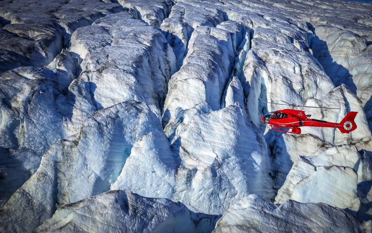 A red helicopter flying low over a broken, deeply crevassed glacier surface